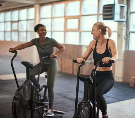 Two fit young women laughing and talking together while riding stationary bikes at the gym