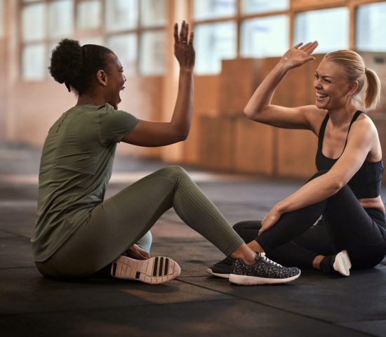 Two fit young women laughing and high-fiving together while sitting on the gym floor after a workout